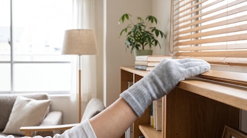 Una mano cubierta con un calcetín gris toca un estante de madera junto a una persiana, libros, una planta y una lámpara.