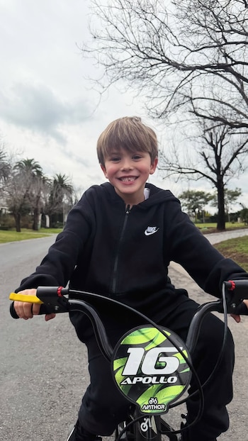 Niño sonriendo en una bicicleta con detalles verdes y negros, viste un buzo y pantalón deportivos negros, fondo con árboles y cielo nublado