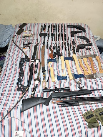 Varias armas, incluyendo pistolas, revólveres, un rifle, katanas, machetes, hachas, cuchillos, un arco, flechas y nunchakus, dispuestas sobre una cama con sábanas a rayas