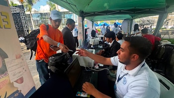 Hombre con camiseta naranja entrega tarjeta a hombre sentado frente a una laptop y una impresora en una mesa. Fondo con carpa verde y personas