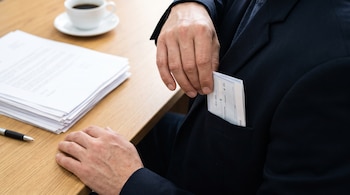 Hombre sin rostro guarda un cheque en el bolsillo interior de su saco negro; en la mesa hay una pila de documentos, un bolígrafo y una taza de café.