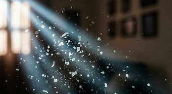 Primer plano de partículas translúcidas y brillantes suspendidas en el aire, iluminadas por haces de luz que cruzan la escena. Al fondo, una ventana y objetos borrosos.