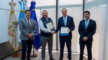 Cuatro hombres de pie; dos estrechan sus manos mientras sostienen documentos. Al fondo se observan tres banderas, una de ellas la de Guatemala y otra con las siglas IGSS