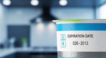Primer plano de un envase de alimento procesado con gotas de condensación. Se lee 'EXPIRATION DATE 026-2013' y un símbolo de refrigeración. Fondo de cocina desenfocado.