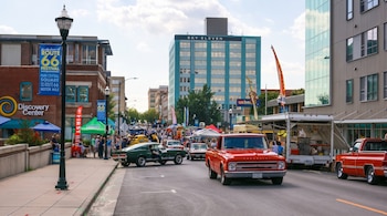 Festival callejero en Springfield con gente, autos clásicos rojos y verdes, carpas, puestos de comida y edificios, incluyendo el 'Sky Eleven', bajo un cielo nublado.