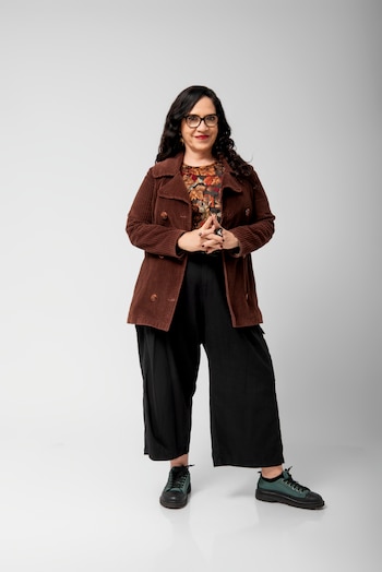 Una mujer de cabello oscuro, gafas, chaqueta de pana marrón, blusa estampada, pantalones negros anchos y zapatos bicolores. Fondo gris claro