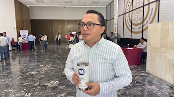 Un hombre con anteojos y camisa de rayas sostiene un vaso térmico en un amplio vestíbulo con piso de mármol y personas al fondo.