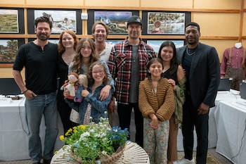 Un grupo diverso de personas, incluyendo adultos y niños sonrientes, posa frente a una pared con obras de arte de temática histórica. Una niña sostiene dos muñecas