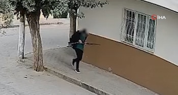 Una persona vestida con chaqueta verde y pantalón negro camina por la acera con un rifle cerca de la fachada de una casa