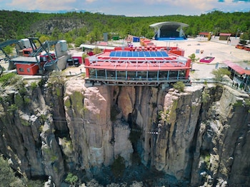 Edificio rojo con paneles solares sobre acantilado rocoso, puente colgante, estación de teleférico, cables, bosque de pinos, escenario al aire libre