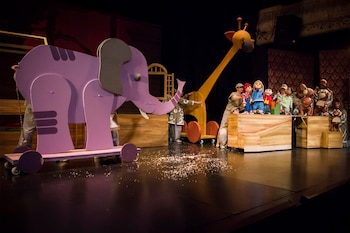 En un escenario, artistas manipulan un gran elefante morado y una jirafa amarilla de madera, junto a muñecos más pequeños. Hay confeti blanco en el suelo
