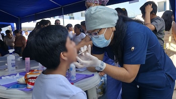 Una mujer con uniforme médico, mascarilla y guantes examina la boca de un joven bajo una carpa azul, con una mesa con productos y un modelo dental