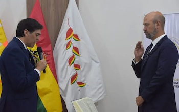 Dos hombres en traje; uno habla por micrófono, el otro con mano derecha levantada. Dos banderas, una roja, amarilla y verde; otra blanca con diseño de hojas. Un libro abierto