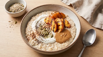 Tazón de avena con yogur, gajos de melocotón asado, crema de almendra y semillas, dispuesto con una cuchara sobre una mesa de madera.