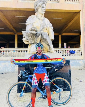 Un ciclista con casco sostiene una bufanda que dice RDC CONGO junto a su bicicleta y frente a una estatua de piedra