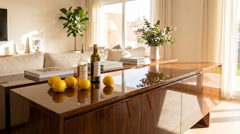 Una sala de estar moderna con un mueble de madera brillante. Sobre él, limones, una botella de vinagre y una de aceite de oliva. Sofá claro y ventana luminosa al fondo.