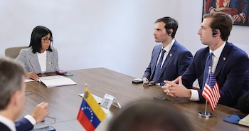Una mujer y dos hombres se sientan en una mesa de madera. Hay banderas de Venezuela y Estados Unidos, documentos y teléfonos