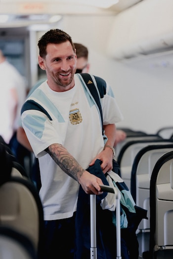 Lionel Messi, con barba, sonríe en el pasillo de un avión. Viste camiseta y mochila de la selección argentina, y tira de una maleta