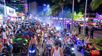 Multitud de motociclistas en una avenida de Acapulco de noche, con motos de colores, luces neón. Patrullas policiales y agentes de seguridad vigilan la escena.