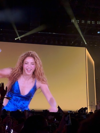 Shakira sonriendo, con cabello largo y rubio, y un top azul, proyectada en una pantalla gigante en un concierto. Una multitud difusa la observa