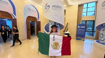 Una persona sonriente con una blusa bordada sostiene la bandera de México, una medalla y un peluche de panda frente a un cartel de IMO 2026 Shanghái, China.