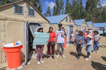 Varias personas permanecen de pie junto a módulos de vivienda prefabricados en un campo, mientras una mujer sostiene un cartel manuscrito