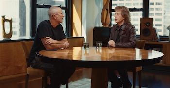 Dos hombres sentados en una mesa de madera. Uno, Mick Jagger, con chaqueta de cuero. El otro, calvo con gafas. Dos vasos de agua y un móvil en la mesa. Ventana y edificios de fondo