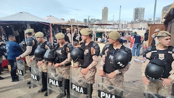Oficiales de Prefectura Naval Argentina en uniforme antidisturbios con cascos y escudos. Están en una zona de puestos de feria con edificios de ciudad al fondo