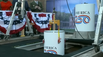 Cápsula del tiempo blanca con logo America 250 siendo bajada por una grúa en un pozo, junto a otra cápsula similar y decoraciones de bandera estadounidense