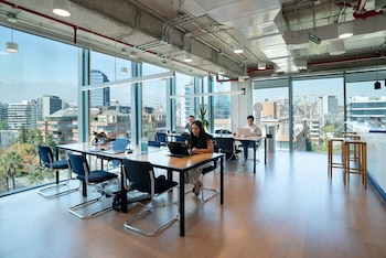 Oficina moderna de planta abierta con grandes ventanales. Varias personas trabajan en mesas compartidas. Sillas, computadoras portátiles y vista a la ciudad