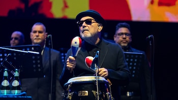 Rubén Blades, con sombrero, gafas de sol y barba, toca maracas de Panamá y un tambor conga en un escenario oscuro con músicos detrás
