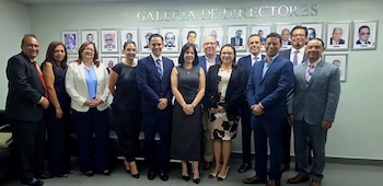 Trece personas, hombres y mujeres, de pie en una oficina. Detrás, una pared blanca con retratos enmarcados y el texto GALERIA DE DIRECTORES