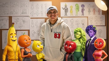 Hombre sonriente con gorra y sudadera, rodeado de siete personajes 3D de frutas. Al fondo, tablero con bocetos y diseños de sus creaciones.
