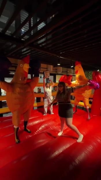 Dos personas con disfraces de pollo y guantes de boxeo sobre un ring inflable rojo. Una mujer con pompones. Otra mujer sujeta un cartel
