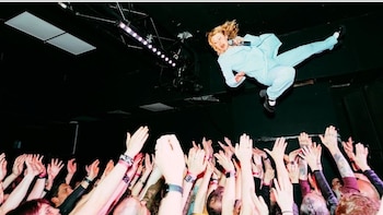 Una cantante vestida con un traje celeste salta por el aire sobre una multitud de personas que levantan las manos en un concierto