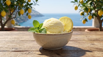 Un tazón de cerámica con una bola de gelato amarillo, una rodaja de limón y menta sobre una mesa de madera ante limoneros y el mar.