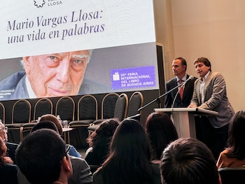 Dos hombres hablan en un atril frente a una pantalla grande con una imagen del rostro de Mario Vargas Llosa y texto sobre la Feria del Libro de Buenos Aires