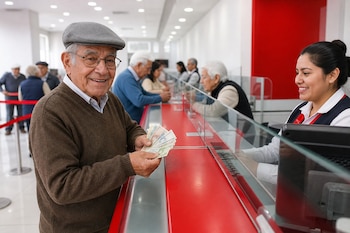 Un hombre adulto mayor sostiene dinero en efectivo en la ventanilla de un banco ante la mirada de una cajera.