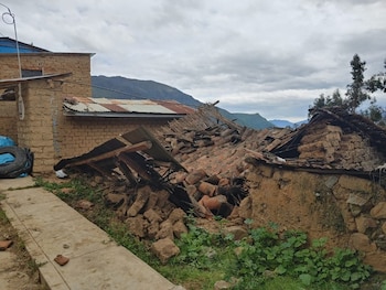 Restos de una vivienda rústica derrumbada, con escombros de adobe, tejas y madera. Se ven paredes parciales, un sendero de cemento y montañas al fondo bajo un cielo nublado
