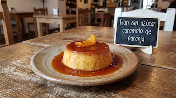 Flan con caramelo y una rodaja de naranja sobre un plato de cerámica, junto a un pequeño cartel de pizarra en una mesa de madera.