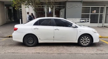 Automóvil sedán blanco estacionado en la calle frente a un edificio. Presenta un neumático trasero de auxilio y rasguños en el lateral