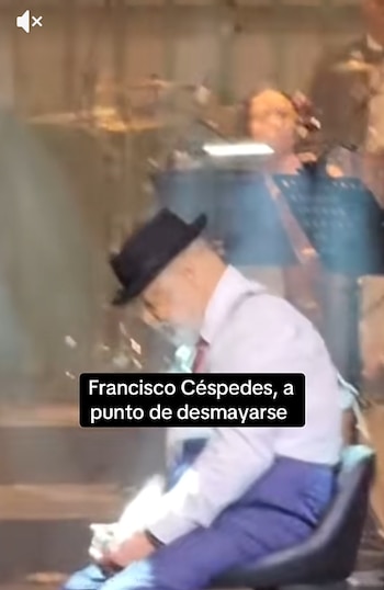 Un hombre con sombrero y camisa clara permanece sentado en una silla con la cabeza inclinada en un escenario junto a músicos