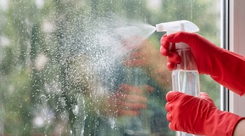 Unas manos con guantes rojos de goma sostienen un frasco pulverizador que rocía líquido blanco sobre un vidrio sucio.
