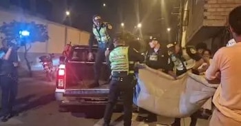 En la noche, policías suben a una persona envuelta en tela clara a la parte trasera de una camioneta pickup. Luces intensas iluminan la escena y se ven espectadores