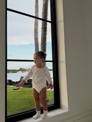 Una niña pequeña, con un body blanco estampado y calcetines blancos, de pie sobre el alféizar de una ventana, con palmeras, agua y césped en el exterior