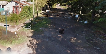 Vista aérea de una calle de tierra con charcos y vegetación densa. A la izquierda, un poste con señal de tránsito 160 y 46. Al fondo, casas modestas y árboles