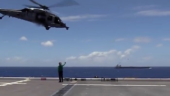 Helicóptero Sikorsky UH-60 Black Hawk gris despegando/aterrizando sobre la cubierta de un buque. Un tripulante en verde señala. Mar azul y otro barco al fondo