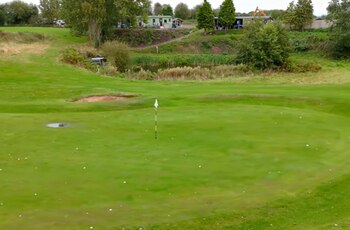 Campo de golf con green verde, bandera blanca, múltiples bolas de golf, bunker de arena, charco de agua y edificaciones con árboles al fondo