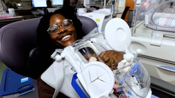 Una mujer con gafas sostiene a un bebé dentro de un contenedor médico transparente en una sala con equipos de monitoreo