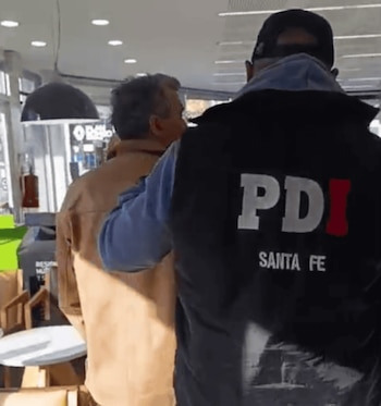 Un hombre de espaldas con camisa marrón es escoltado por un agente de PDI Santa Fe, que viste chaleco oscuro y gorra. Se observa el interior de un local